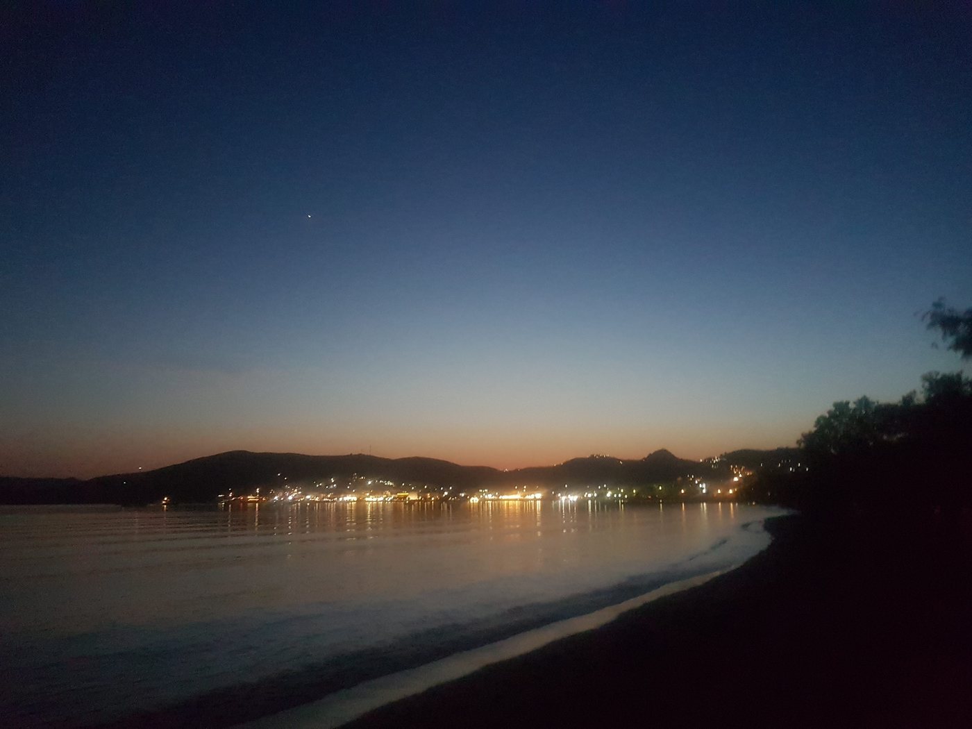 The port town of Adamas lit up at dusk around its wide curving bay on Milos
