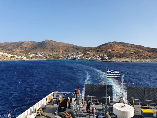 The stern deck of a Greek ferry underway, flag flying over the wake with a Cycladic island village receding in the distance