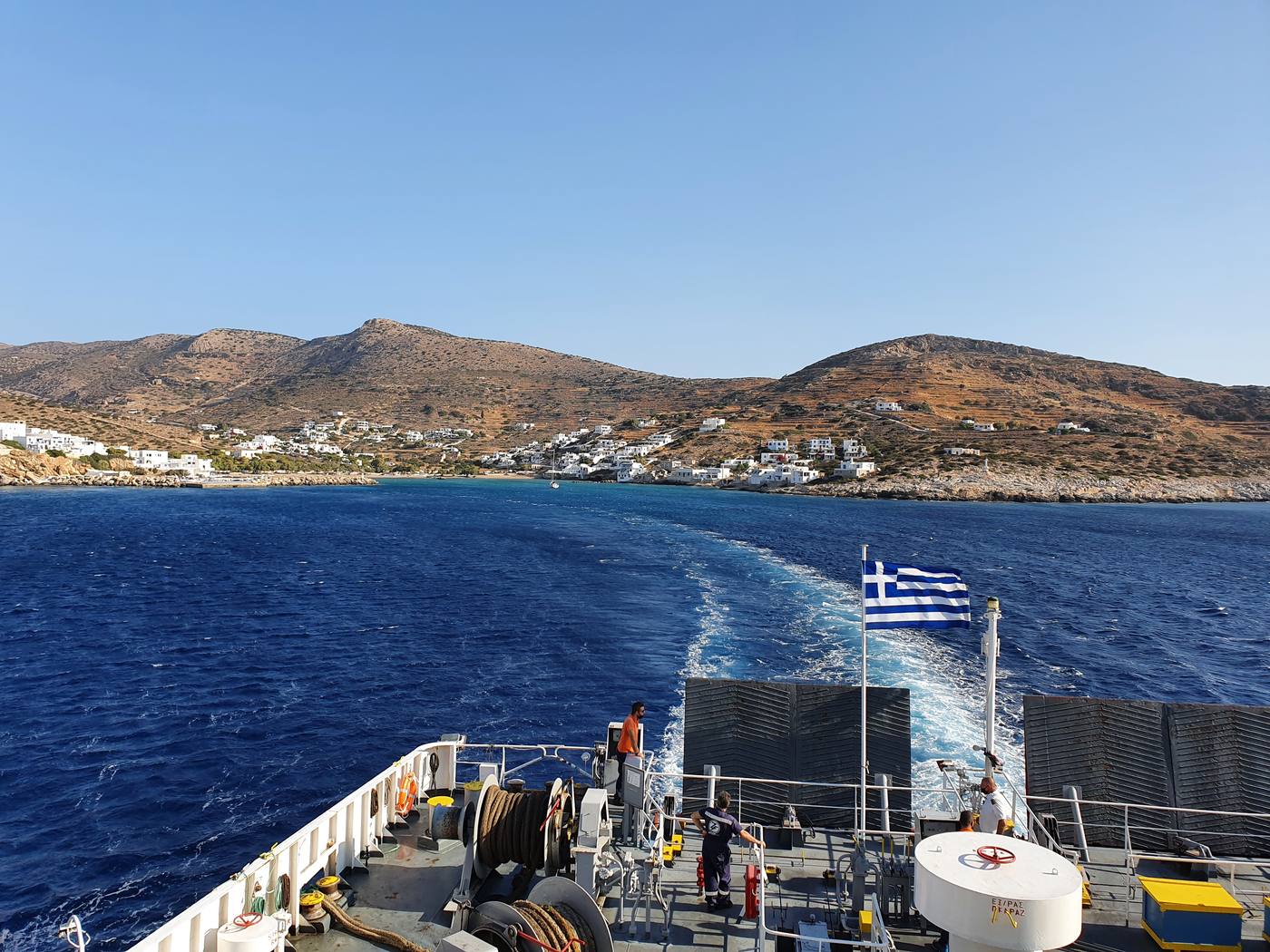 The stern deck of a Greek ferry underway, flag flying over the wake with a Cycladic island village receding in the distance