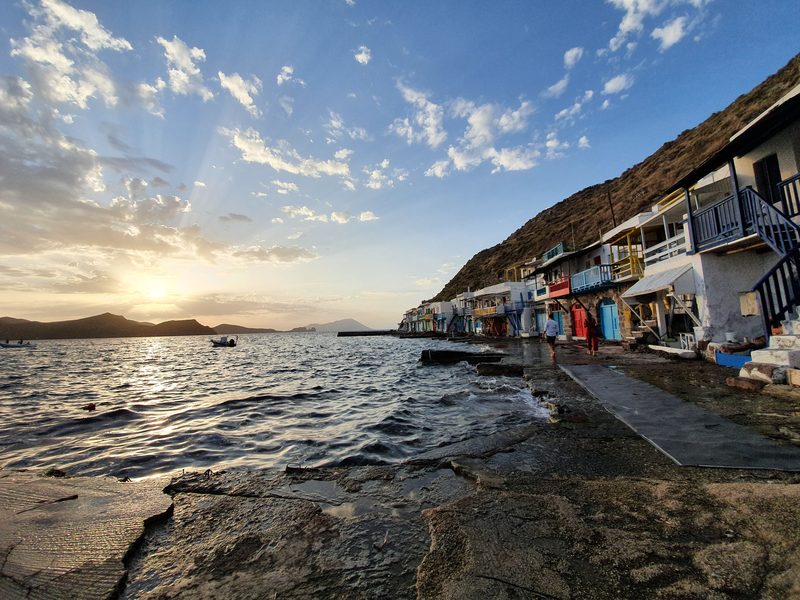 Visitors walking the narrow waterside path in front of Klima's painted syrmata at sunset