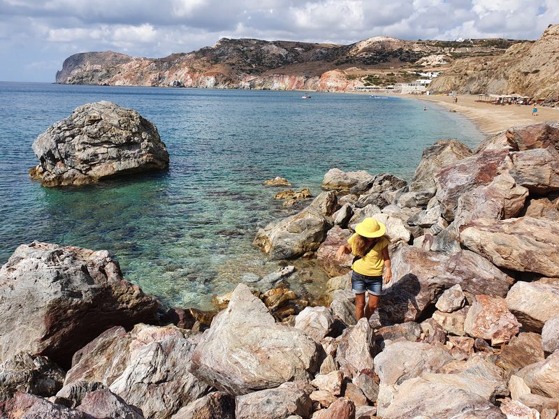 A walker scrambling over large volcanic boulders above the sea at Paleochori, with red and ochre cliffs beyond