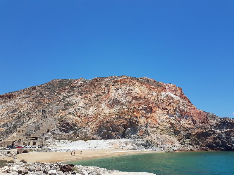 The tiered stone buildings of the old Paliorema sulphur works on Milos, set against a red and white mineral-streaked cliff