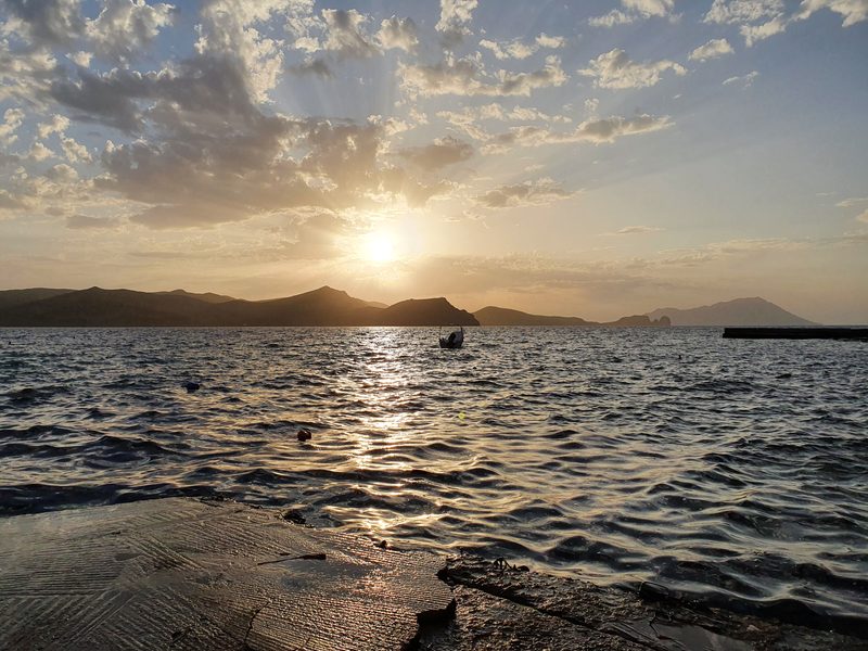The sun breaking through cloud over the sea off the west coast of Milos, with a small fishing boat silhouetted on the water