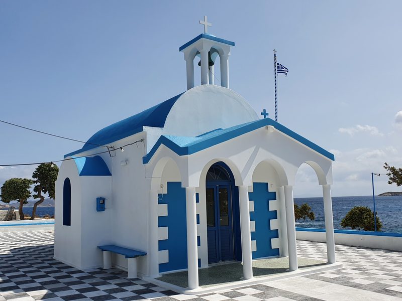 A small blue-domed chapel on the seafront point at Pollonia, with the bay and hills behind