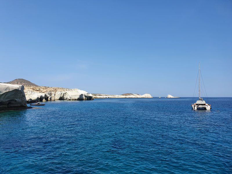 A catamaran anchored off the white headlands near Sarakiniko, with a small boat closer to shore
