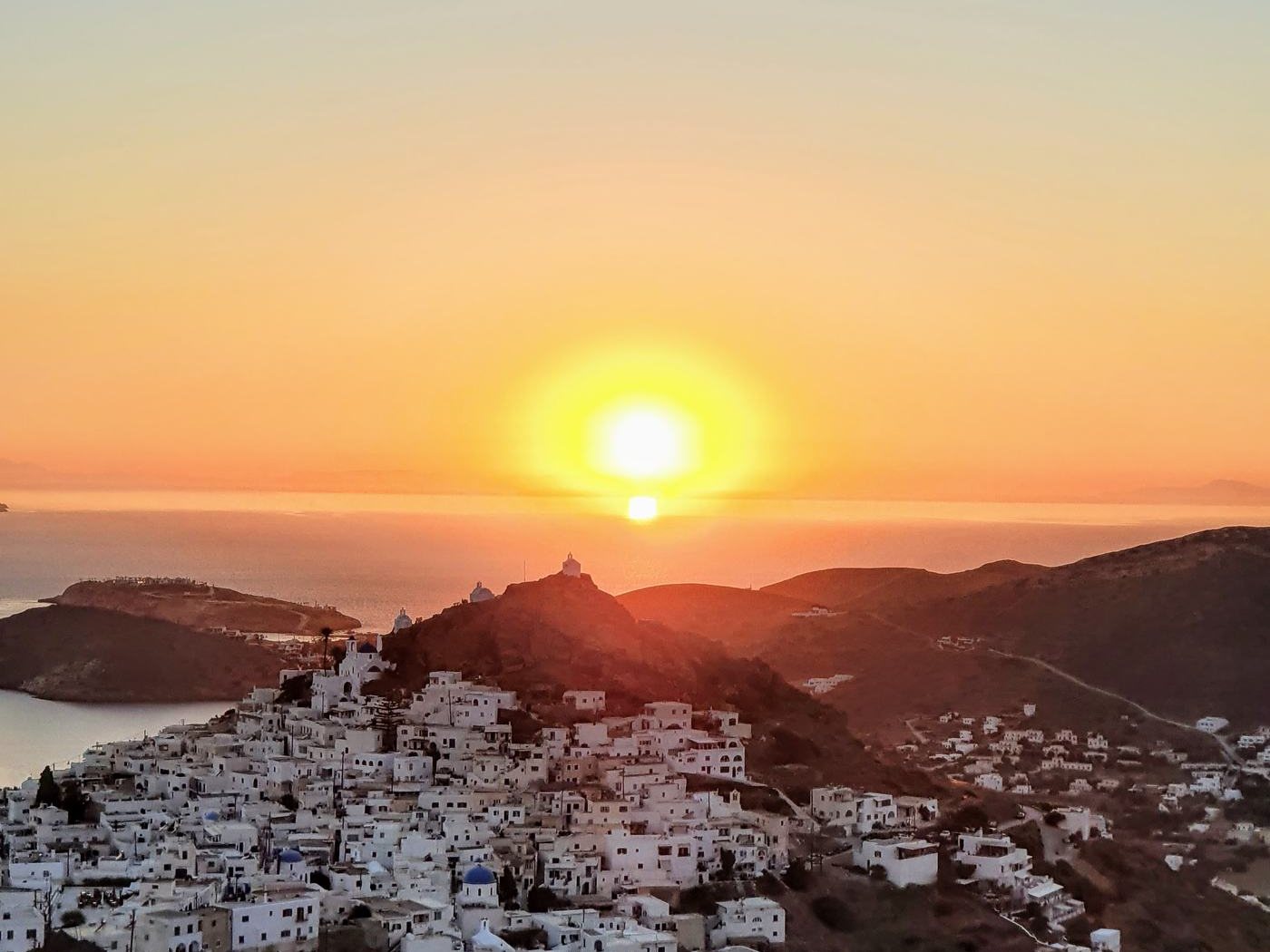 Sunset over Ios Chora, whitewashed houses on the hillside with a windmill silhouette against the glowing sky