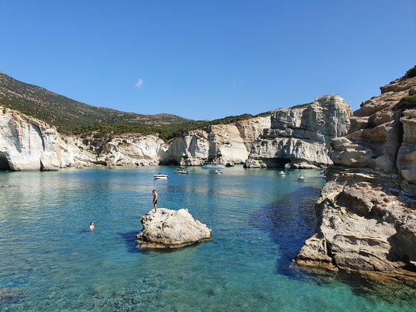 Towering white sea stacks with cave arches rise from turquoise water at Kleftiko, a swimmer standing on a rock in the foreground