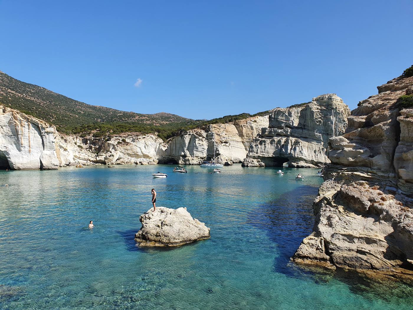 Towering white sea stacks with cave arches rise from turquoise water at Kleftiko, a swimmer standing on a rock in the foreground
