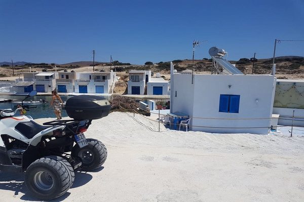 A rented ATV parked beside a small fishing harbour in Milos, with whitewashed boathouses behind