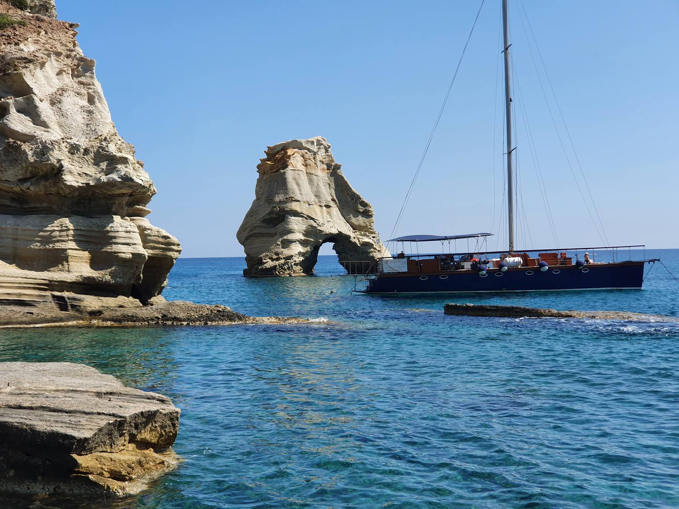 A Milos boat tour vessel anchored beside a tall sea-arch rock stack, with a swimmer in the clear water alongside
