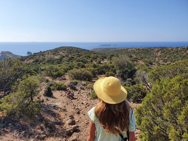 A hiker on a dirt trail across a scrubby headland on the Milos coast, with the sea and small islets beyond