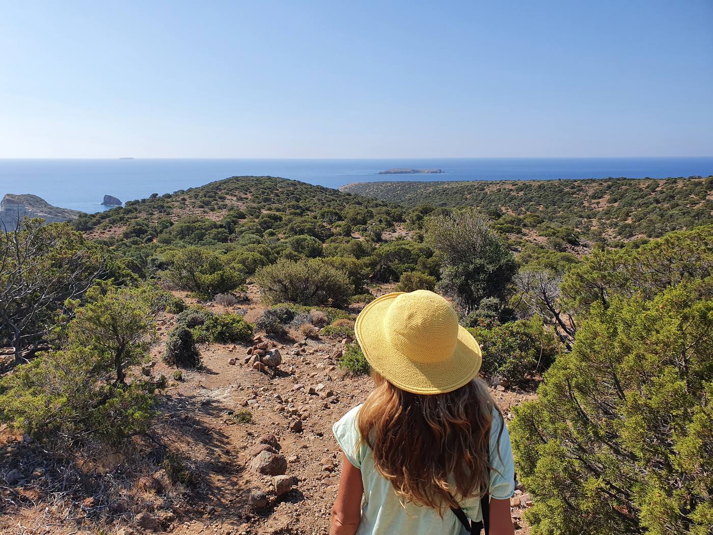 A hiker on a dirt trail across a scrubby headland on the Milos coast, with the sea and small islets beyond