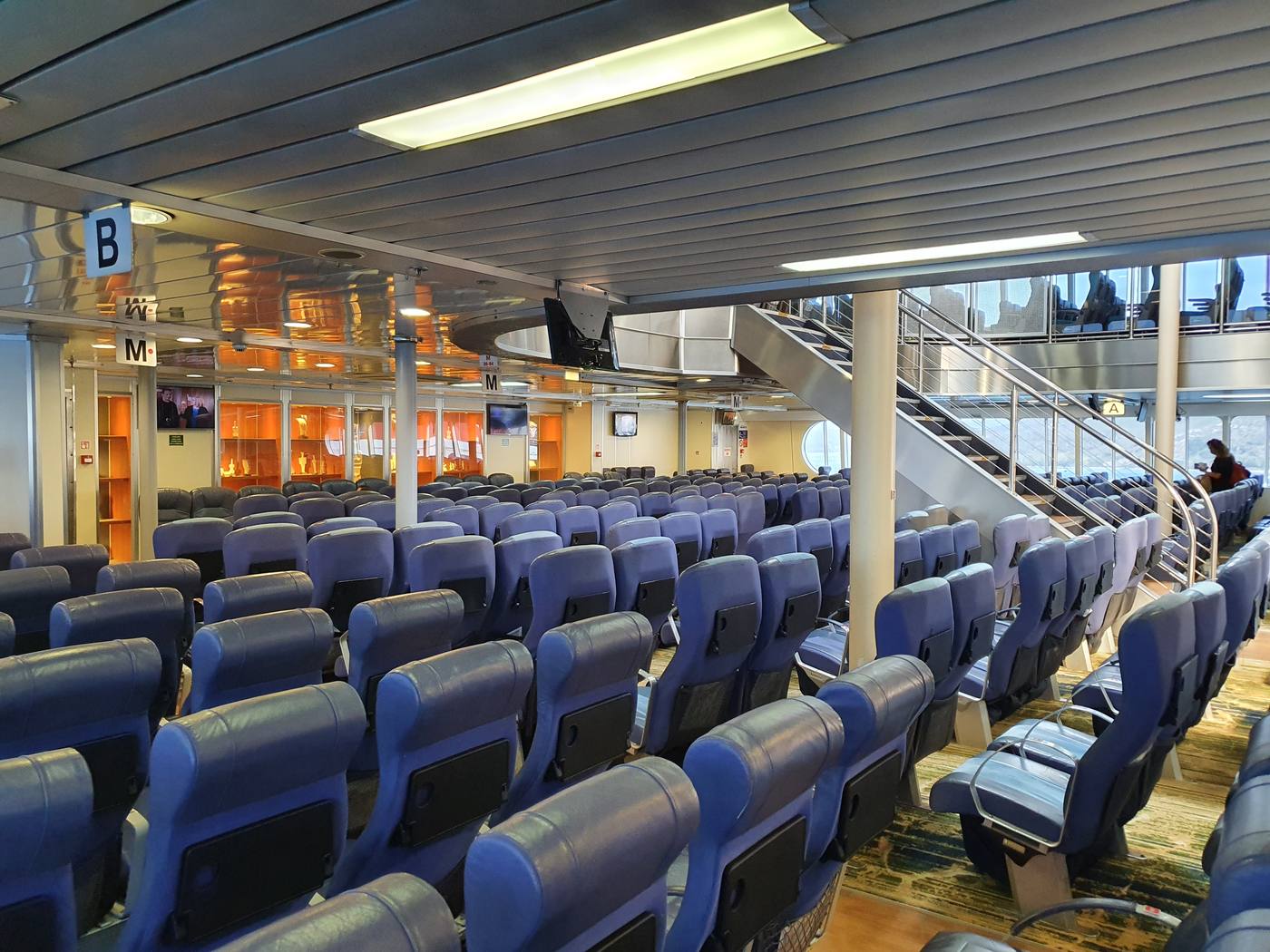 Rows of passenger seats aboard a Greek ferry, with a staircase leading up to the open deck