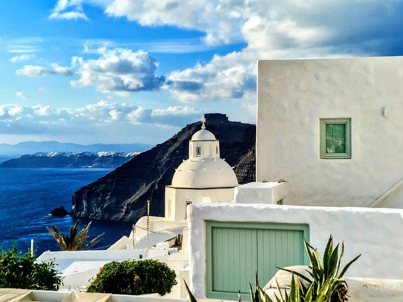 A blue-shuttered church dome above the Santorini caldera, whitewashed walls against the sea below