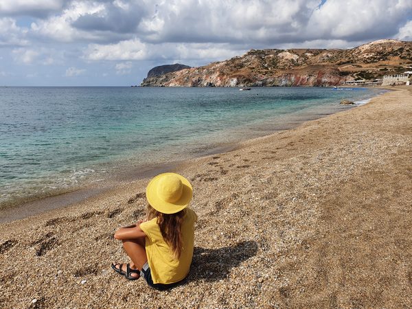 A visitor sits on the sand at Paleochori beach, facing the sea, with the striped red and ochre cliffs of the bay in the distance