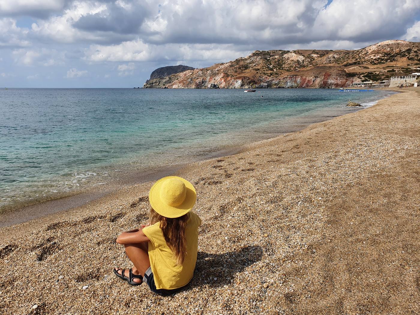 A visitor sits on the sand at Paleochori beach, facing the sea, with the striped red and ochre cliffs of the bay in the distance