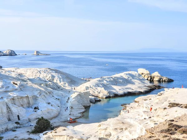 A wide view over Sarakiniko's white lunar rock formations and the narrow turquoise inlet, with sea stacks visible offshore